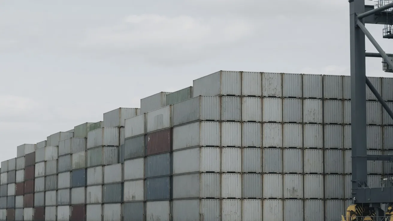 Stacked containers at a deep sea terminal under an overcast sky, with the leg of a quay crane at the right of the frame.